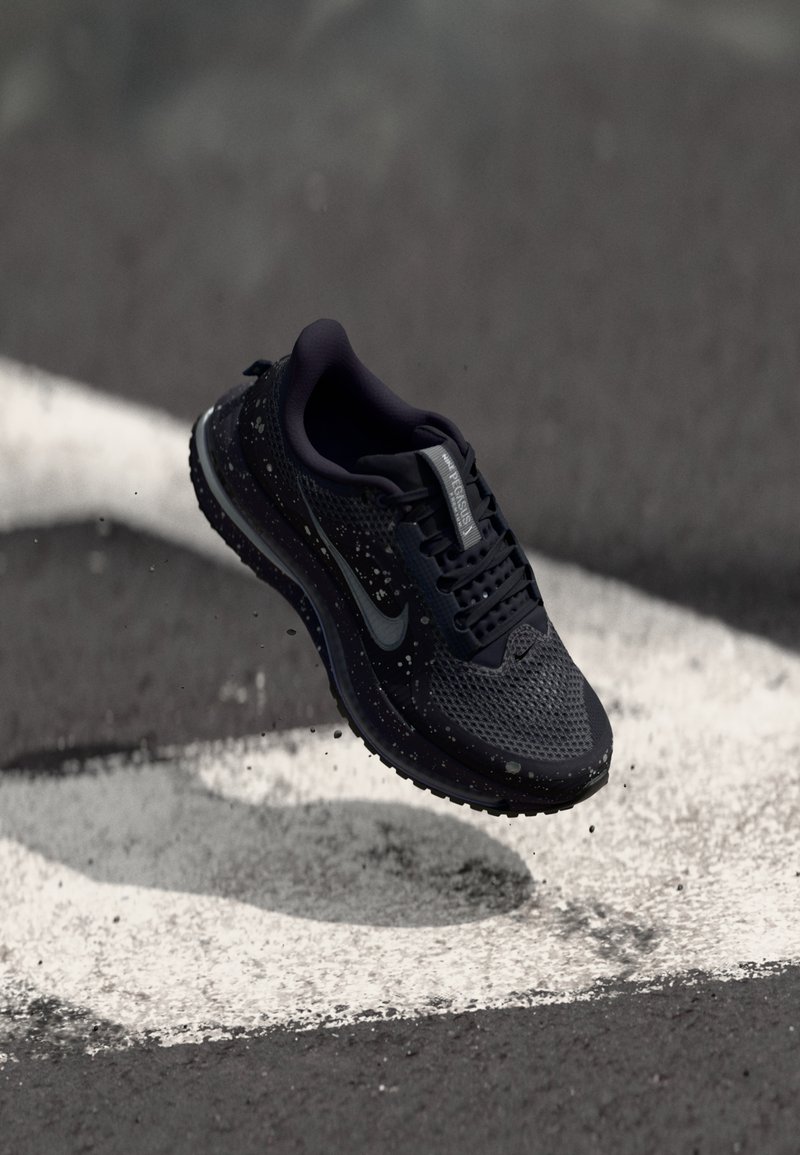 Nike teka za tek v temno modri barvi s zračnim mrežastim zgornjim delom, teksturiranim podplatom in odsevnimi poudarki. Vključuje podrobnosti s kapljanjem barve.