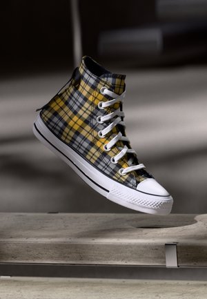 Baskets montantes avec motif à carreaux jaunes et noirs, bout, semelle et lacets blancs, suspendues au-dessus d’un banc en béton.