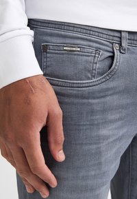 Jeans din denim gri, cu o croială slim, având o buzunar frontal, textură uzată și un etichetă metalică cu brand. O mână vizibilă se odihnește pe buzunar.