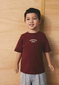 Bordo bomba majica s kratkimi rokavi in belim napisom "Neomejen nasmeh vsak dan," kombinirana s svetlo modrimi denim kratkimi hlačami.