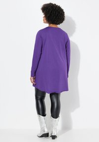 Lila Langarmbluse mit gekurvtem Saum, kombiniert mit schwarzen Kunstlederleggings und silbernen Cowboyboots. Glatte Stofftextur.