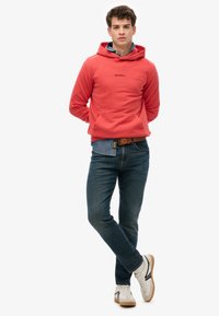 Sudadera roja con capucha y bolsillo tipo canguro, logotipo en el pecho. Combinada con vaqueros oscuros y zapatillas blancas con suelas marrones, en una pose de pie.