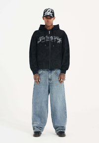 Black zip-up hoodie with hood, featuring gray ornate text design. Paired with light blue wide-leg jeans and black shoes, standing against a white background.