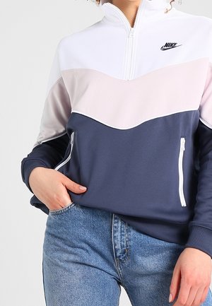 Henkilö pukeutuneena väriblokki-Nike-vetoketjutakkiin, jossa on valkoinen, vaaleanpunainen ja laivastonsininen sävy sekä vetoketjulliset taskut, yhdistettynä sinisiin farkkuihin.