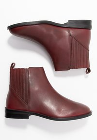 Un par de botas de cuero burdeos suaves hasta el tobillo con suelas negras, paneles elásticos en los laterales y lengüetas en un fondo blanco.