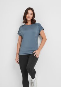 Woman in a blue short-sleeve athletic shirt, black leggings, and white sneakers standing with one leg bent against a white wall.