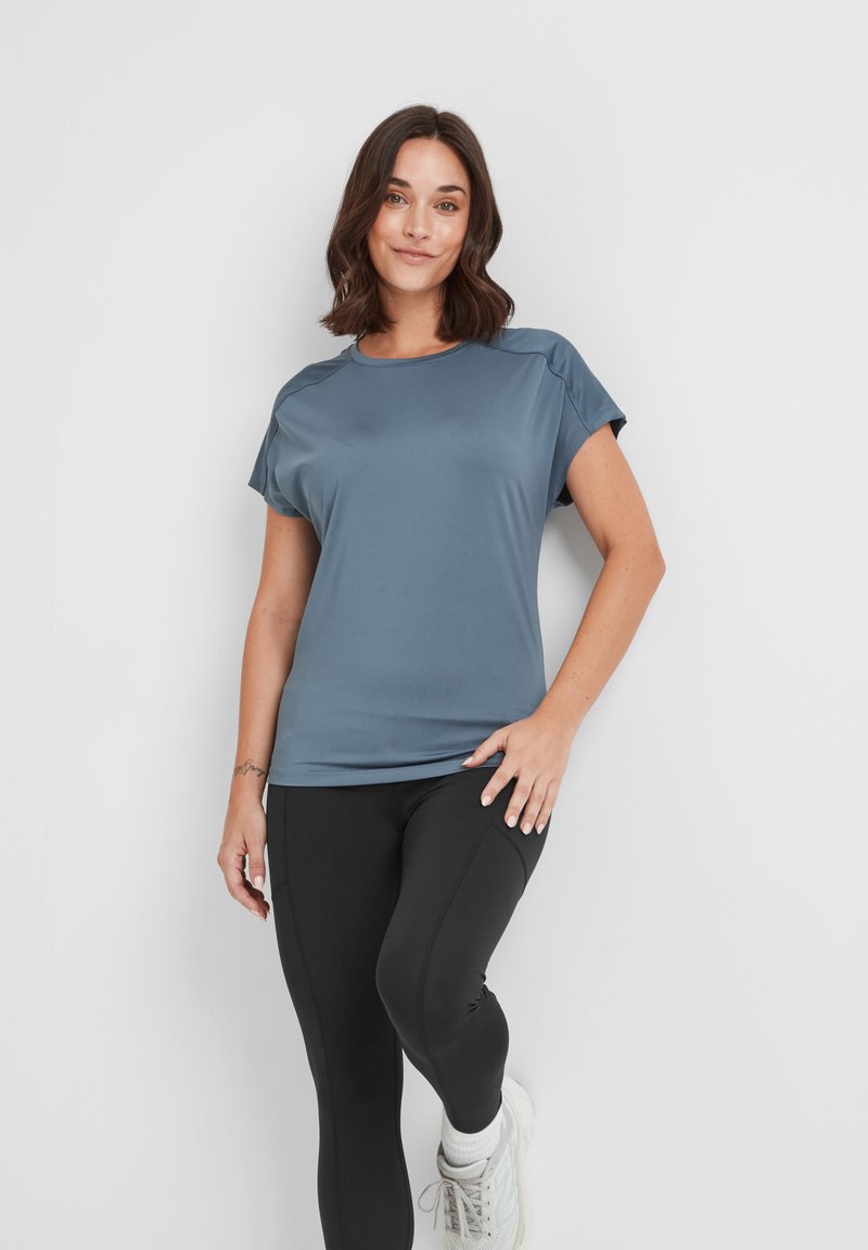 Woman in a blue short-sleeve athletic shirt, black leggings, and white sneakers standing with one leg bent against a white wall.