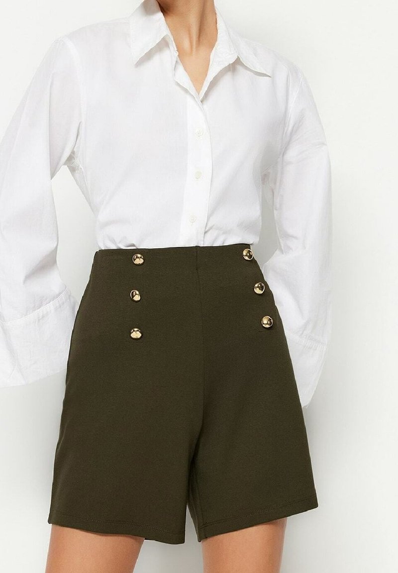 Femme portant un short taille haute vert olive avec six boutons dorés associé à une chemise blanche à manches longues boutonnée.