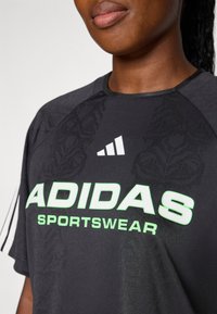 Juodas sportinis marškinėliai su tekstūriniu raštu, su užrašu "ADIDAS" neoninės žalios spalvos ir "SPORTSWEAR" po juo, akcentuotas baltais trimis juostomis ant rankovių.