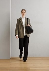 Blazer oversize verde oliva, camicia bianca con colletto, pantaloni neri e scarpe nere lucide. Borsa nera con tracolla. Ambientazione minimalista.