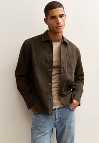 Camisa verde oliva de botones con un bolsillo en el pecho, llevada sobre una camiseta color beige claro, combinada con unos jeans azul claro. Textura de tela suave.