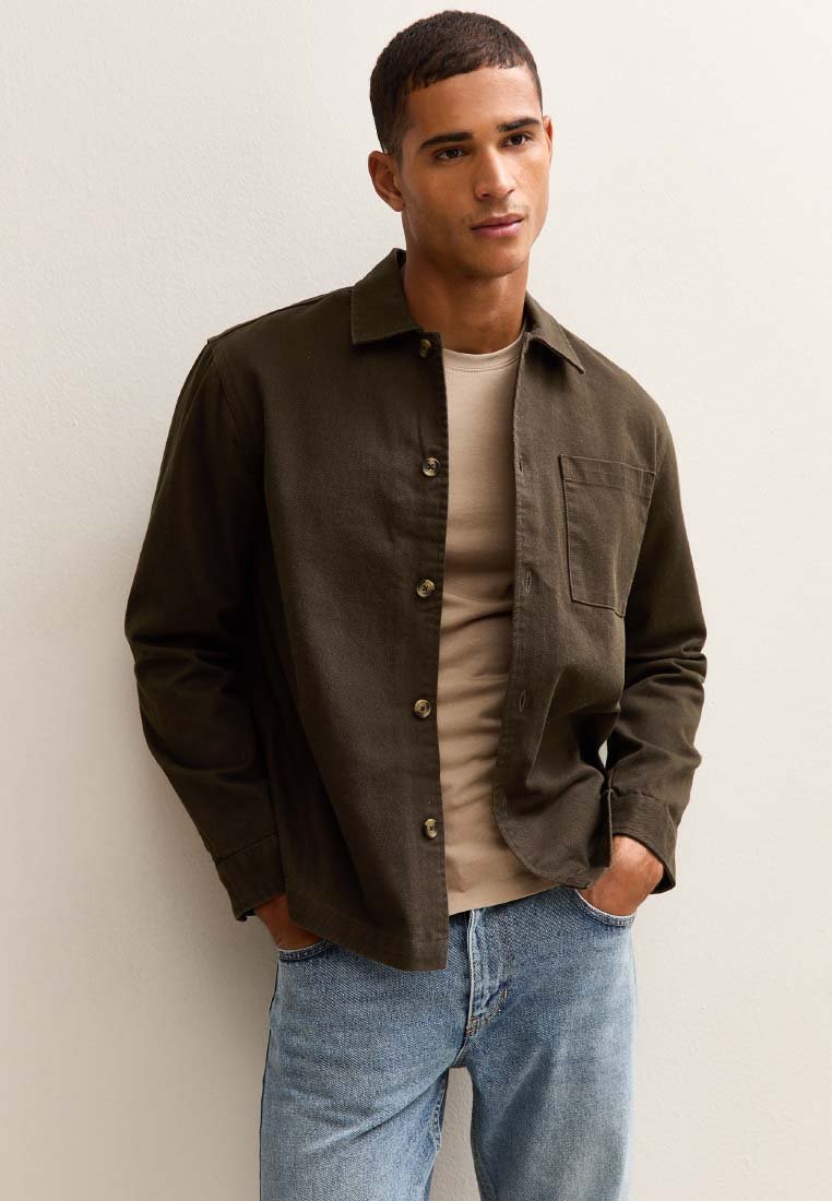 Camisa verde oliva de botones con un bolsillo en el pecho, llevada sobre una camiseta color beige claro, combinada con unos jeans azul claro. Textura de tela suave.