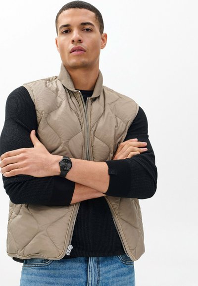 Mężczyzna z założonymi skrzyżowanymi ramionami, ubrany w beżową pikowaną kamizelkę, czarny teksturowany sweter, niebieskie dżinsy i czarny zegarek na rękę, na jednolitym tle.