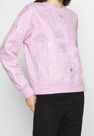 Lichtpaarse trui met goudomrande Nike-logo's, de tekst "Just do it" en bloemenpatronen, gedragen door een persoon in een zwarte broek.
