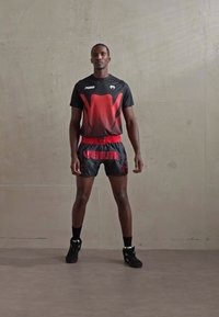Athlet steht an einer schlichten Wand und trägt ein schwarzes und rotes Sportoutfit, einschließlich Shorts mit der Aufschrift "VENUM" und passenden Sportschuhen.
