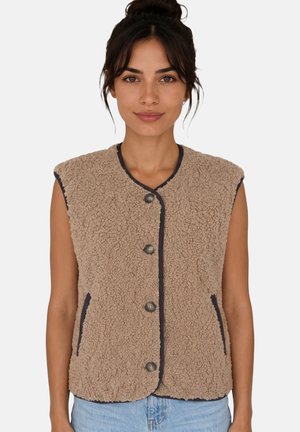 Mujer con chaleco sherpa sin mangas de color beige con botones sobre una camiseta negra y vaqueros azul claro, con el pelo recogido en un moño despeinado.