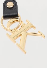 Μπρελόκ από μέταλλο σε χρυσό τόνο με διαπλεκόμενα γράμματα "T" και "K", προσαρτημένο σε μαύρο δερμάτινο λουράκι με στρογγυλό χρυσό κουμπί.