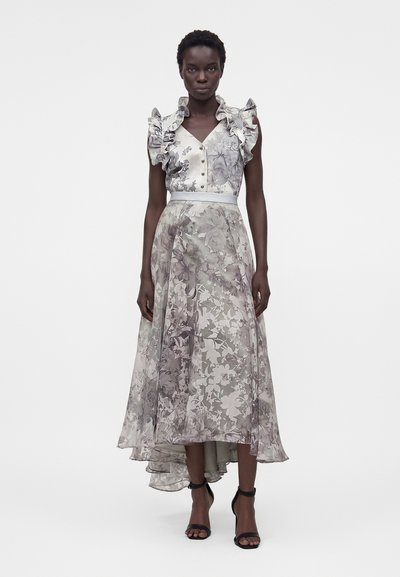 Γυναίκα φοράει ένα αμάνικο, λουλουδάτο, μίντι φόρεμα με βολάν, κουμπιά και ασύμμετρο τελείωμα, σε συνδυασμό με μαύρες γόβες με λουράκι στον αστράγαλο.