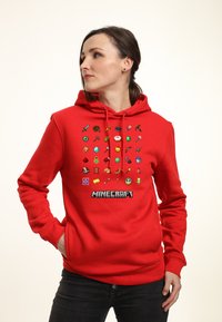Czerwona bluza z kapturem z grafiką na przodzie przedstawiającą 24 pikselowe ikony związane z Minecraftem oraz słowo "MINECRAFT" napisane pogrubioną czarną czcionką.
