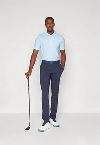 Ljusblå pikétröja med krage, matchad med mörkblå byxor och vita golfskor. Står med en golfklubba.