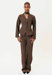 Hnedý prispôsobený oblek s priliehavou bundou s hlbokým výstrihom do V a detailom šatky, doplnený o rovné nohavice a špicaté čierne topánky.