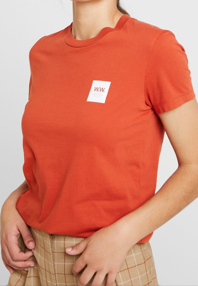 Tričko s krátkým rukávem a volným střihem v jasně oranžové barvě z bavlny, s malým bílým čtvercovým logem s "W.W." na levé části hrudi.