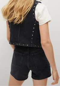 Chaleco de mezclilla negro con tachuelas plateadas, combinado con un bodysuit negro y shorts negros. La tela es texturada y ajustada, con bolsillos traseros.