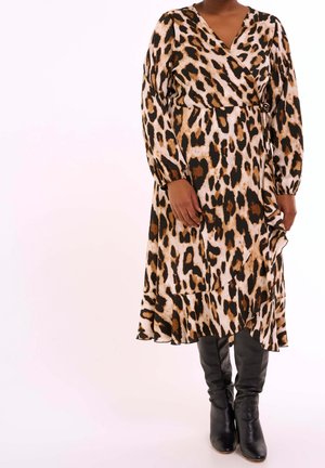 Vrouw draagt een langermige wikkeljurk met luipaardprint, gecombineerd met zwarte leren kniehoge laarzen, staand tegen een lichte achtergrond.