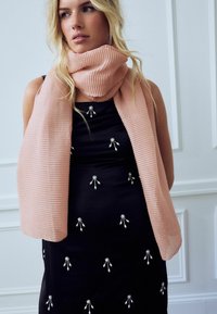 Écharpe côtelée rose clair enroulée autour du cou, associée à une robe noire avec des détails de broche en argent. Tissu lisse avec des accents texturés.