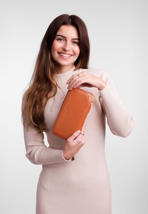 Brown leather wallet with a cork accent. Rectangular shape, zipper closure, embossed logo. Model holds the wallet, wearing a ribbed beige dress.