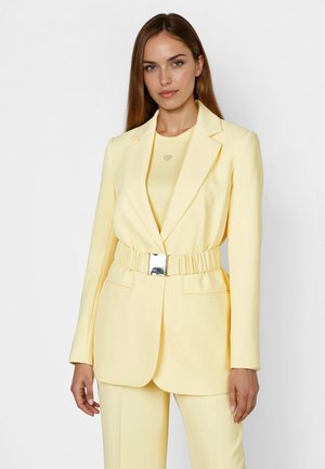 Frau mit langem braunem Haar, die ein blassgelbes, tailliertes Blazer und passende Hose trägt, vor einem schlichten weißen Hintergrund steht.