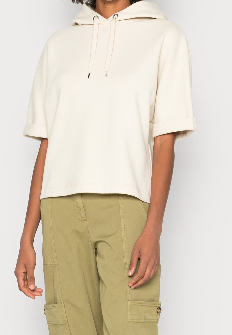 Femme portant un sweat à capuche crème à manches courtes avec des cordons et un pantalon cargo kaki, debout devant un fond uni.