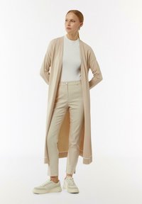 Langer, offener Cardigan aus weichem beigem Strick. Kombiniert mit engen beigen Hosen und weißen Sneakers. Glatte Textur mit minimalistischem Design.