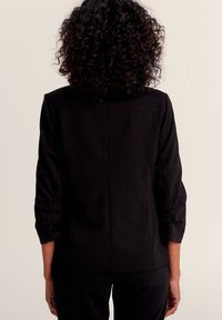 Veste noire avec des manches trois-quarts, design structuré, tissu lisse, présentant une texture subtile et une couture arrière droite.