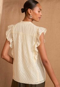 Cream-colored blouse with ruffled sleeves, featuring a wave pattern and sheer texture. Back view highlights the gathered seam and soft drape.
