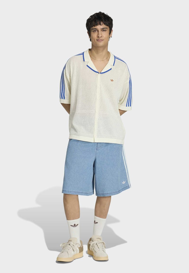 Jonge man die met zijn handen achter zijn rug staat, gekleed in een wit mesh shirt met blauwe strepen, lichtblauwe knielange korte broek, witte sokken en beige sneakers.