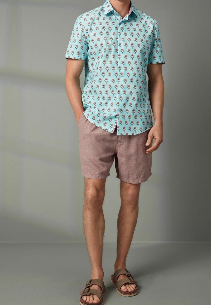 Homme portant une chemise bleu clair à manches courtes avec un motif floral, un short mauve et des sandales marron, debout avec une main dans la poche.