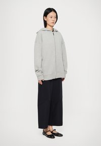 Grijze zip-up hoodie van zacht materiaal, met een capuchon en een oversized pasvorm, gecombineerd met losse donkere broeken en zwarte schoenen met accenten.