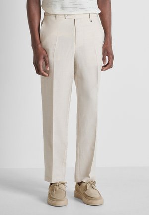 Mand iført beige skræddersyede bukser og matchende beige afslappede sko, stående mod en ensfarvet lys baggrund.