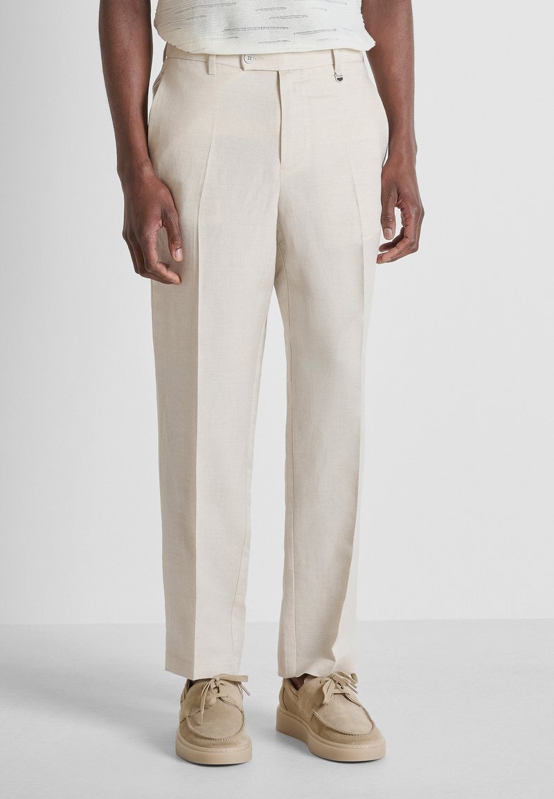 Hombre con pantalones beige a medida y zapatos casuales beige a juego, de pie frente a un fondo claro liso.