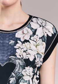 Top con estampado floral en tejido oscuro, que presenta una sección texturizada en azul y un borde plateado en el escote. Mangas cortas y cuello redondeado.