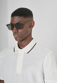 Polo bianco con una finitura testurizzata, dettagli in colletto blu navy e logo ricamato. Occhiali da sole neri quadrati con un design elegante.