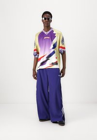 Színes Adidas mez, villámmintával sárga, lila és piros színben. Széles kék nadrággal és fekete cipővel párosítva.