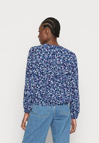 Blusa floral com um fundo azul-marinho e pequenas flores brancas, rosas e verdes. Mangas longas com punhos elásticos e detalhes franzidos.