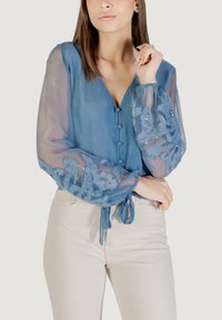 Blusa azul, transparente, con mangas largas y abullonadas que presenta patrones florales bordados y lentejuelas; incluye un cierre frontal con botones y cintas en la cintura.