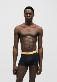 Άνδρας μοντέλο στέκεται μπροστά σε μονόχρωμο φόντο φορώντας μαύρα μπόξερ Nike με κίτρινη μέση, κοιτάζοντας μπροστά με ουδέτερη έκφραση.