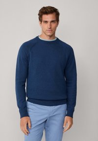 Námornícky modrý sveter z textúrovanej pleteniny, s kruhovým výstrihom a rebrovanými manžetami a spodným okrajom. Nosený cez svetlomodrú košeľu.