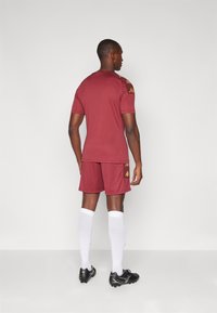 Conjunto de camiseta y pantalones cortos deportivos en color marrón. Fabricado en tejido suave, con detalles en dorado en los hombros y los lados. Botas de fútbol negras y calcetines blancos.