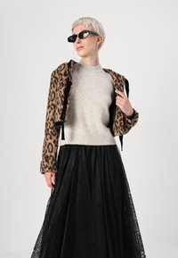 Casaco de padrão de leopardo com cortes, com detalhes em preto, combinado com um suéter cinza claro adornado com pérolas e uma saia maxi de renda preta.