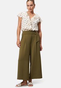 White blouse with floral pattern and ruffle detailing paired with green wide-leg trousers featuring a tied belt accent. Flat sandals complete the look.